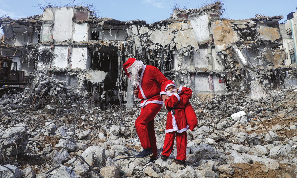 A Palestinian dressed as Santa Claus walks with his child past the al-Jawhara Tower in Gaza City's al-Rimal neighborhood, destroyed by Israeli air strikes last May on Christmas Eve on December 24, 2021. Photo: AFP