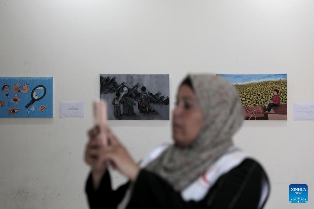 A Palestinian woman takes photos of artworks at a World Mental Health Day exhibition in the southern Gaza Strip city of Khan Younis, on Oct. 10, 2024. A total of 48 Palestinian children who received psychological treatment participated in the exhibition with their paintings. (Photo: Xinhua)