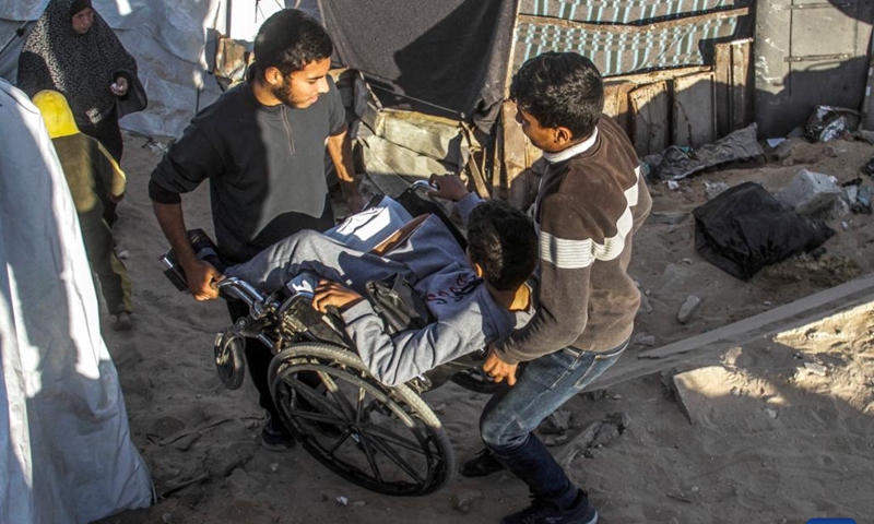 Two men help carry the wheelchair for Akram Harb in Gaza City, on Dec. 13, 2024. Harb, a 13-year-old Palestinian, was injured in an Israeli attack at his house in the Jabalia refugee camp in the northern Gaza Strip in October, 2024. (Photo: Xinhua)